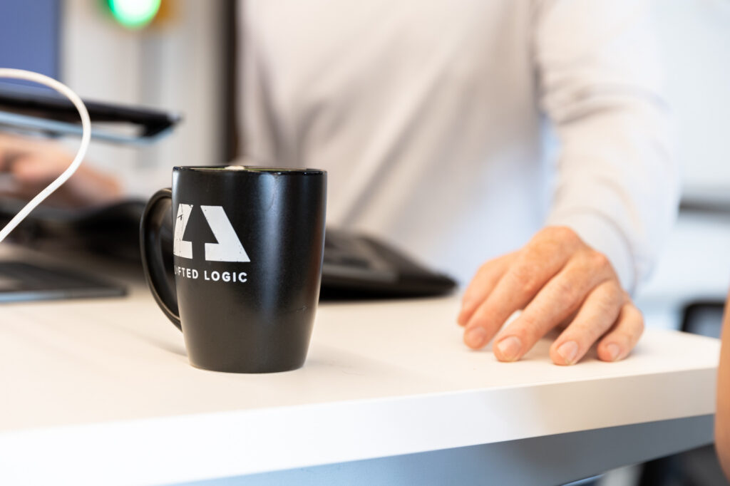Closeup of a Lifted Logic employee's hand next to a branded coffee cup. He's part of the team that supports medical spa owners and managers, include helping them when they're updating their med spa prices.