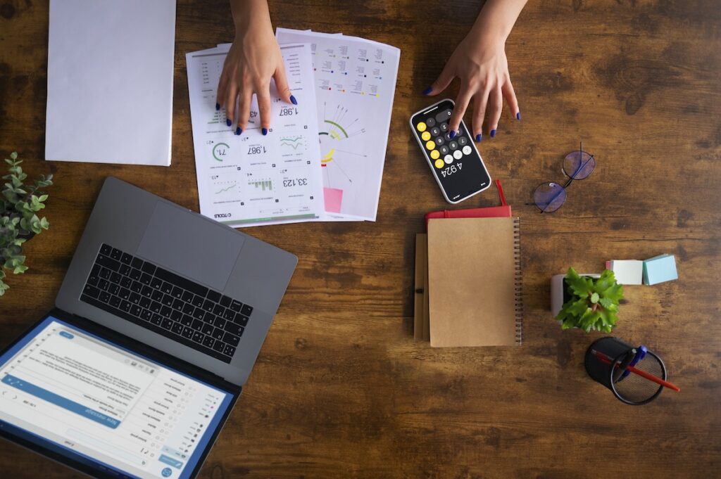 An above view of a woman's hands as she uses a calculator to calculate costs of goods sold. Paper notes and a computer are also visible. At her medical spa she is updating their med spa prices.